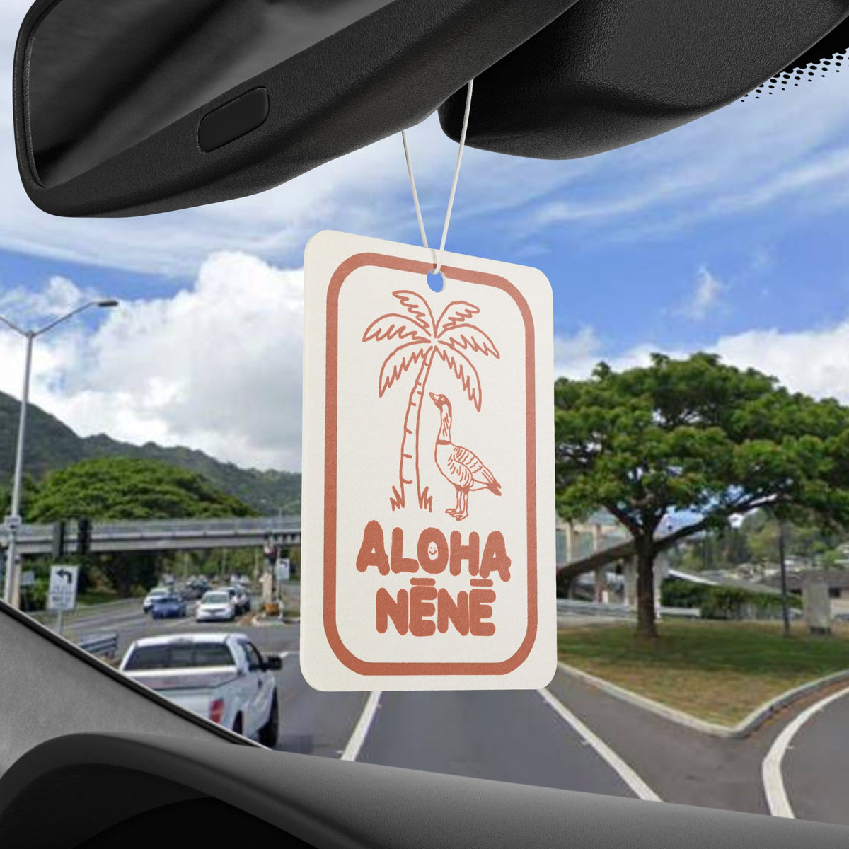 A rectangular car air freshener hanging from the windshield wiper arm inside a vehicle. The white tag has a coral-orange border, a palm tree and goose illustration, and the text 'ALOHA NENE'.