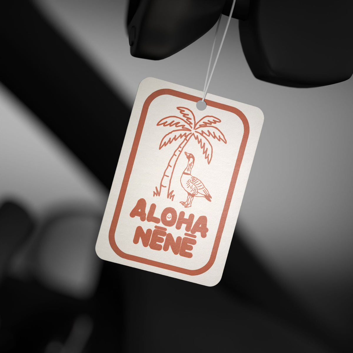A rectangular car air freshener tag with rounded corners, featuring a orange outline illustration of a palm tree and a bird with the words 'ALOHA NENE' printed below. The tag is shown hanging from a black car seat against a blurred black and gray background.