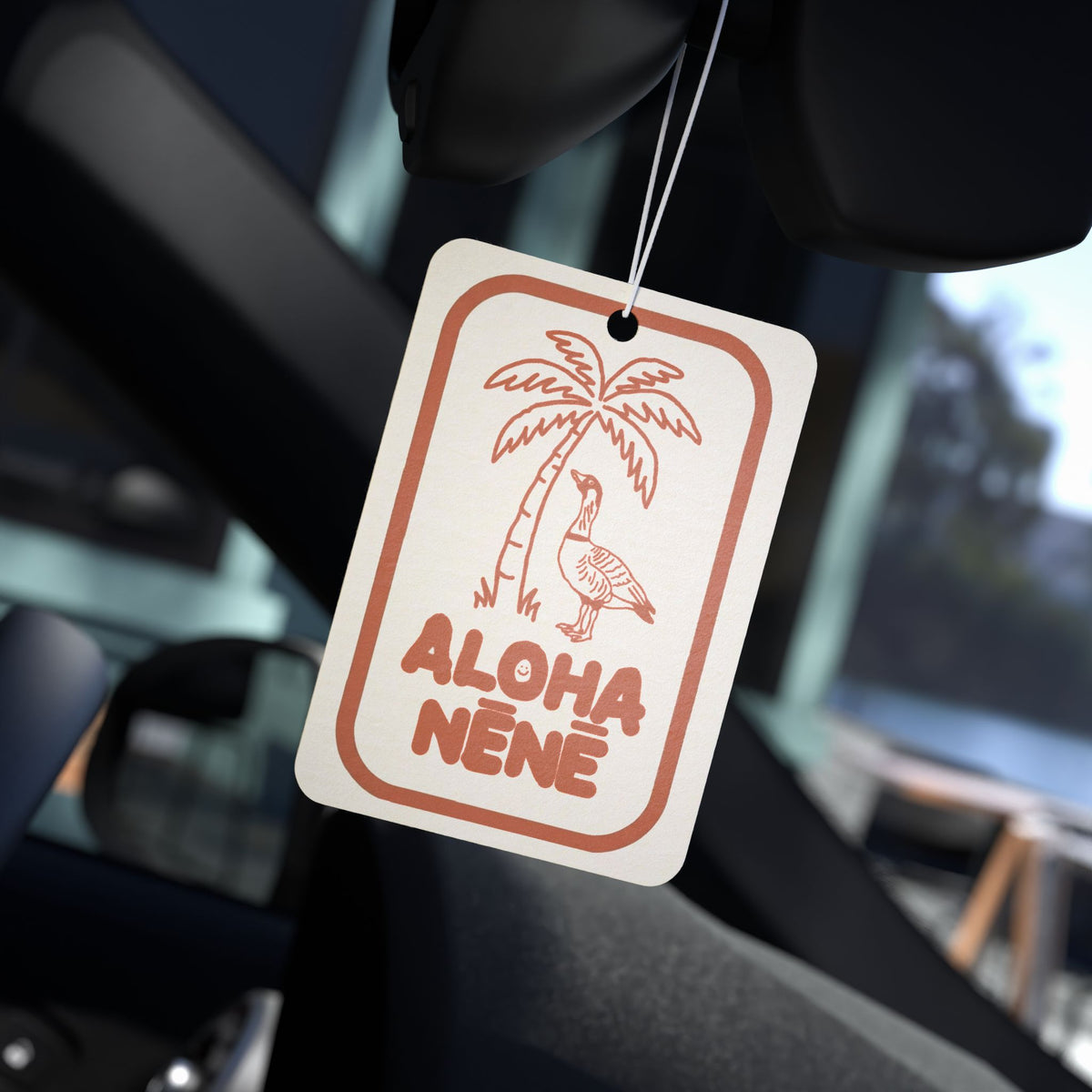 A rectangular car air freshener tag hanging from a windshield wiper inside a vehicle. The tag is off-white with a terracotta orange border and graphic of a palm tree and a bird, with the text 'ALOHA NENE' printed below.