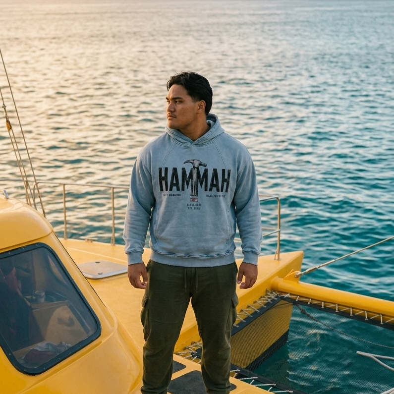 A man wearing a light blue 'HAMMAH' hoodie, dark green cargo pants, and white sneakers stands on the yellow deck of a boat with the ocean water in the background, AlohaNene