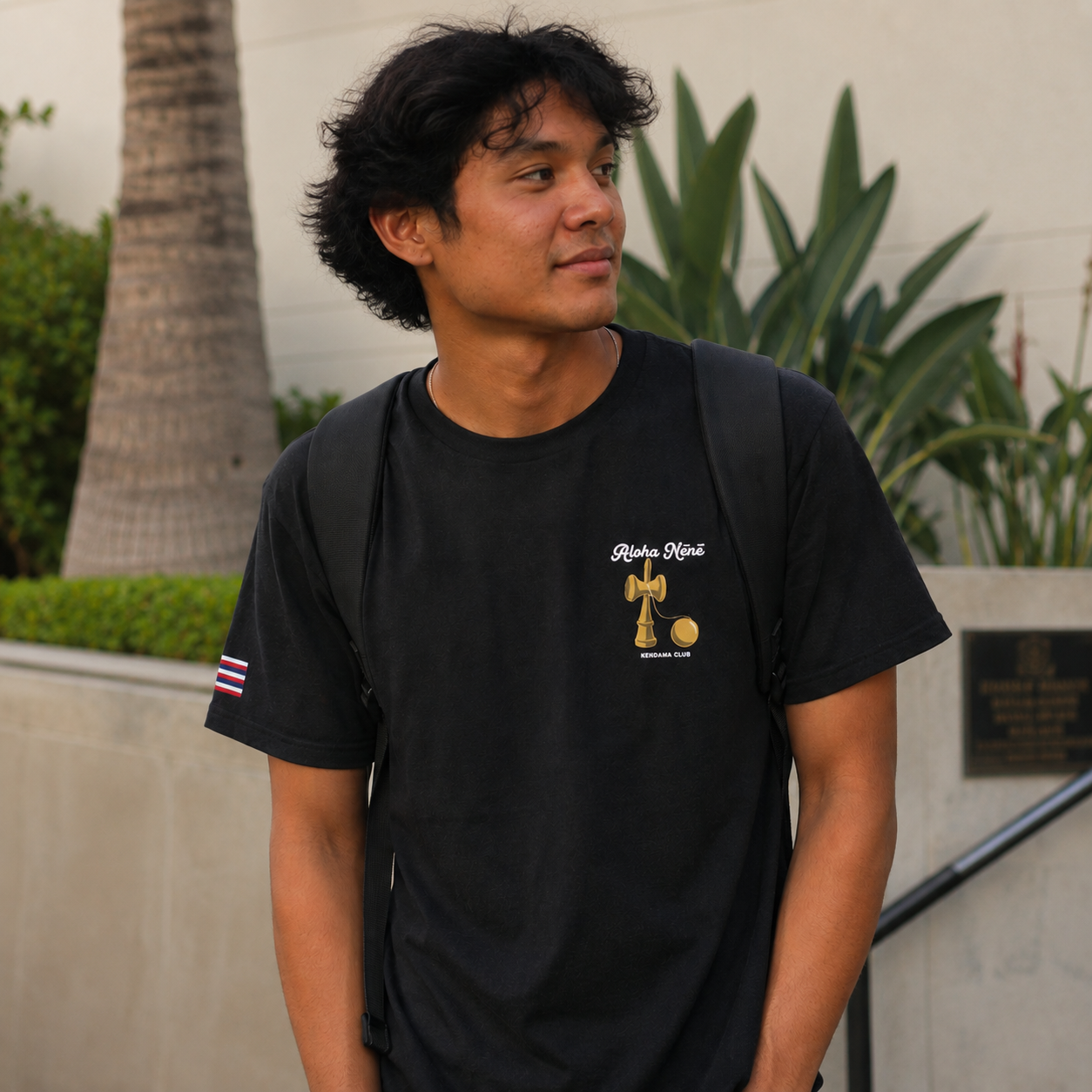 A man wearing a black short-sleeve t-shirt with a gold and white graphic on the left chest and a small Hawaiian flag on the sleeve, standing outdoors in front of a beige wall and green plants.