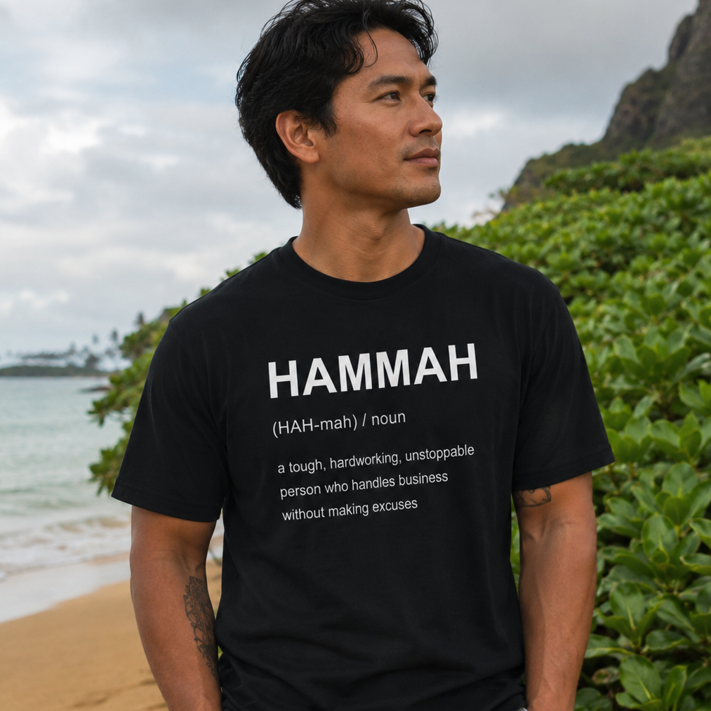 A man wearing a black graphic t-shirt with the word 'HAMMAH' printed in white, standing on a beach with green foliage and ocean in the background.