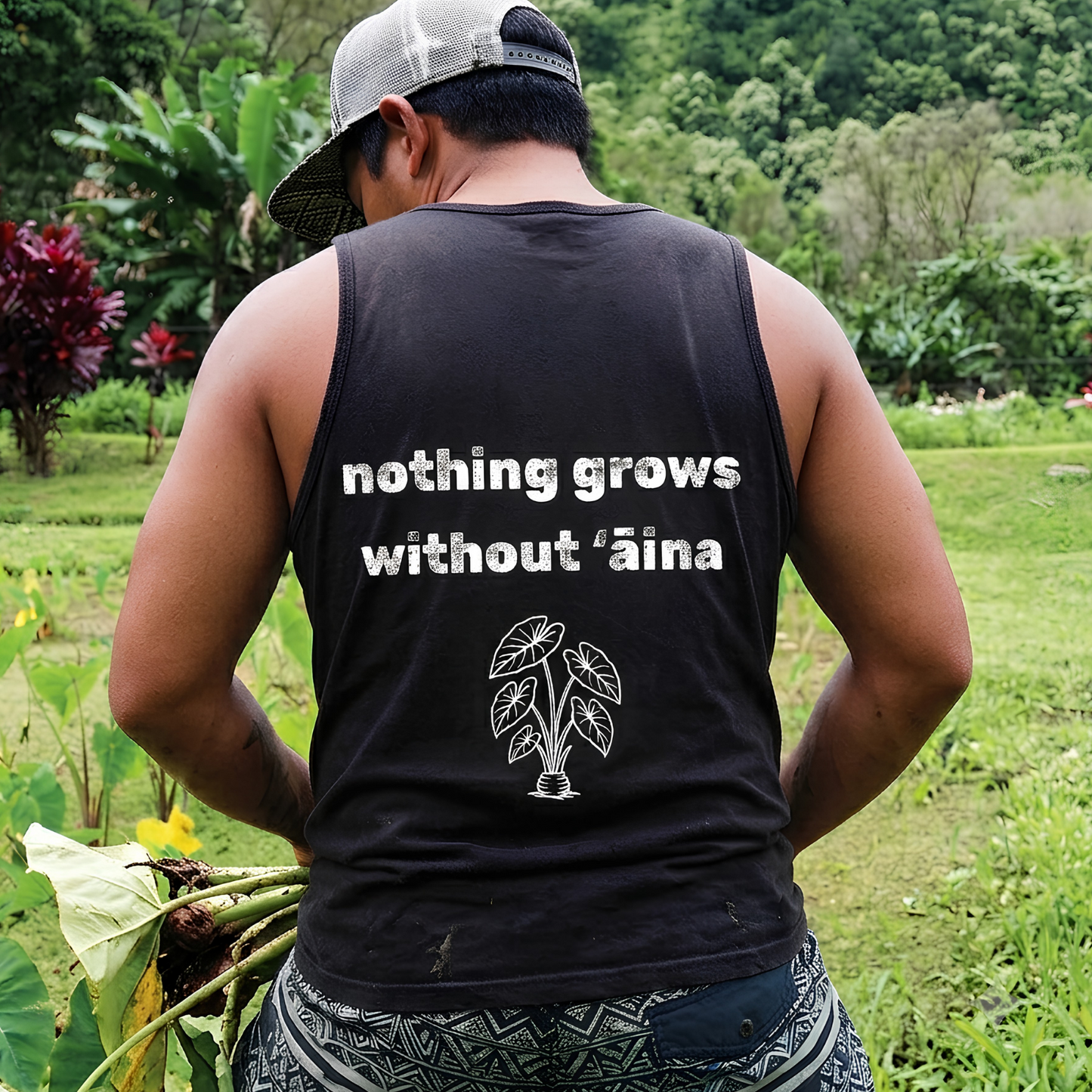 A man viewed from behind wearing a black sleeveless tank top with the white printed text 'nothing grows without ’aina' and a small leaf graphic, standing in a lush green outdoor farm setting.