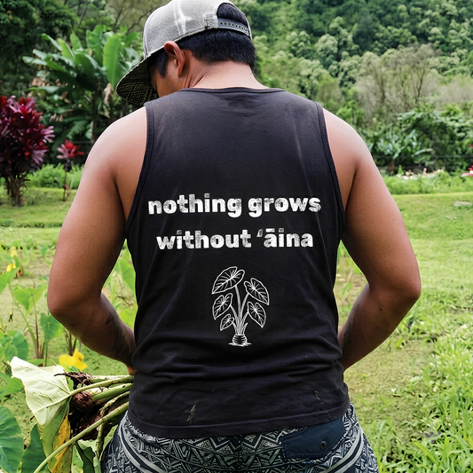 A man viewed from behind wearing a black sleeveless tank top with the white printed text 'nothing grows without ’aina' and a small leaf graphic, standing in a lush green outdoor farm setting.