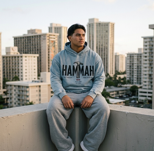 A man sits on a concrete ledge in Waikiki, Hawaii, wearing a light blue hoodie with 'HAMMAH' printed across the chest and matching light gray sweatpants, Aloha Nene and Hawaiian Flag