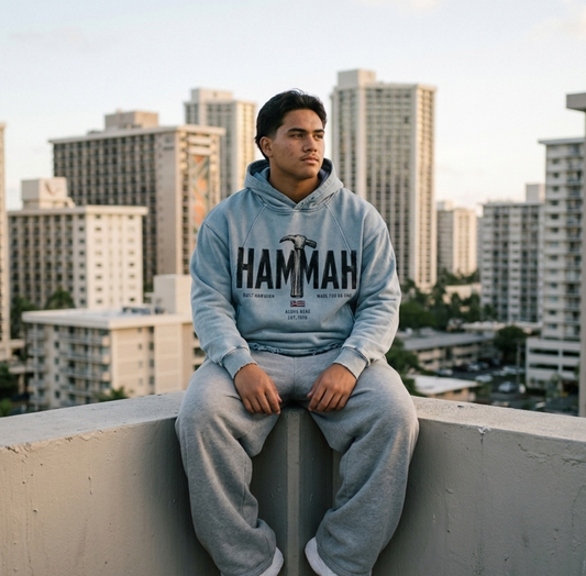 A man sits on a concrete ledge in Waikiki, Hawaii, wearing a light blue hoodie with 'HAMMAH' printed across the chest and matching light gray sweatpants, Aloha Nene and Hawaiian Flag