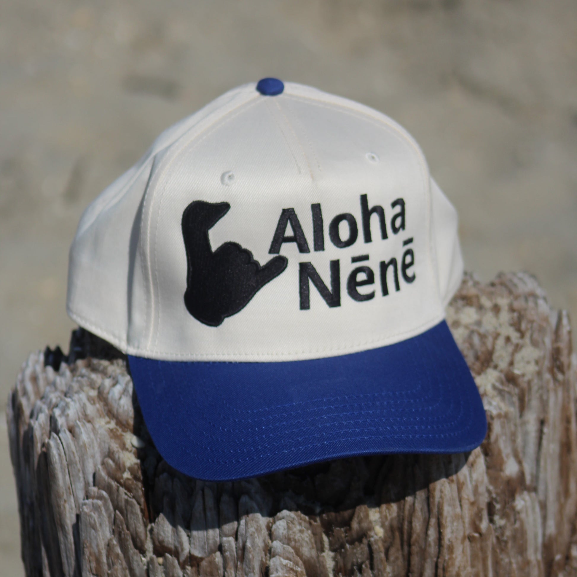 A white and royal blue baseball cap with a black 'Aloha Néné' hand gesture graphic, resting on a weathered wooden post with a sandy beach background.